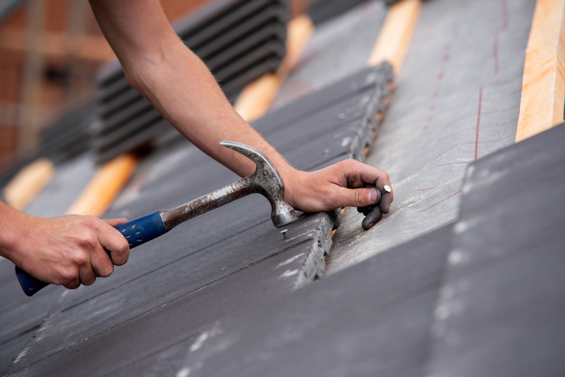 Person hammering a nail into a gray roof tile during roof construction. Person hammering a nail into a gray roof tile during roof construction.