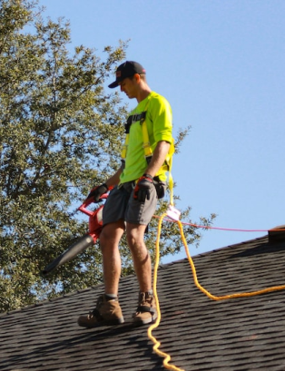 Man on a roof using a blower, wearing safety harness and neon shirt; sunny day.