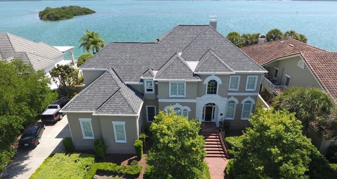 Two-story grey house with a dark grey roof and brick pathway next to water, trees, and other homes. Two-story grey house with a dark grey roof and brick pathway next to water, trees, and other homes.
