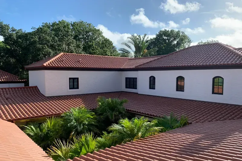 Tiled rooftops of a white building with black windows, surrounding a courtyard filled with tropical plants under a cloudy sky. Tiled rooftops of a white building with black windows, surrounding a courtyard filled with tropical plants under a cloudy sky.