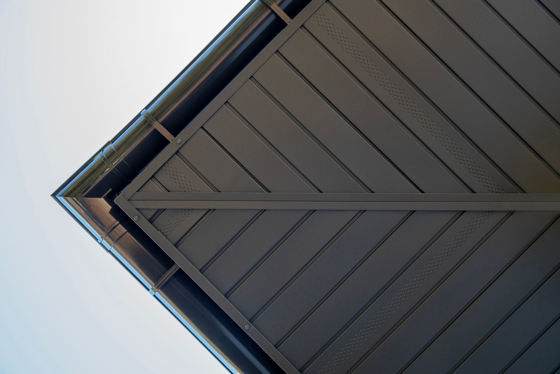 Low-angle view of a gray roof's underside, showing angled wooden planks and a gutter against a light sky.