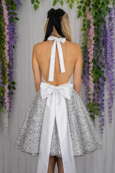 Back view of a person in a white halter dress with a bow, standing before a floral backdrop.