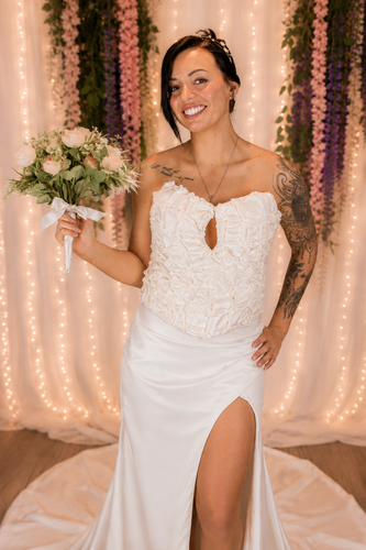 Bride in a white strapless gown holding a bouquet, standing before a floral backdrop and string lights.