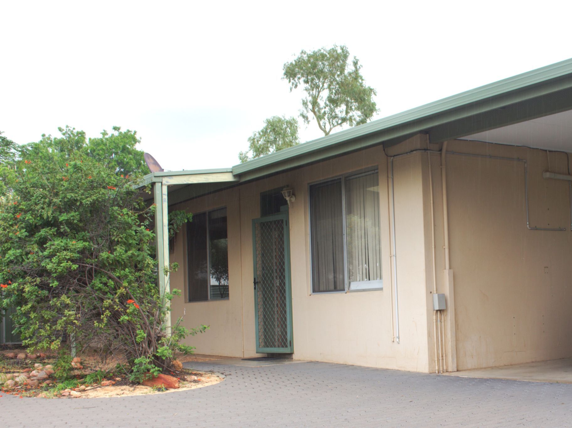 A single-story tan house with a green roof, a covered carport, and a small front garden with bushes on a paved driveway.