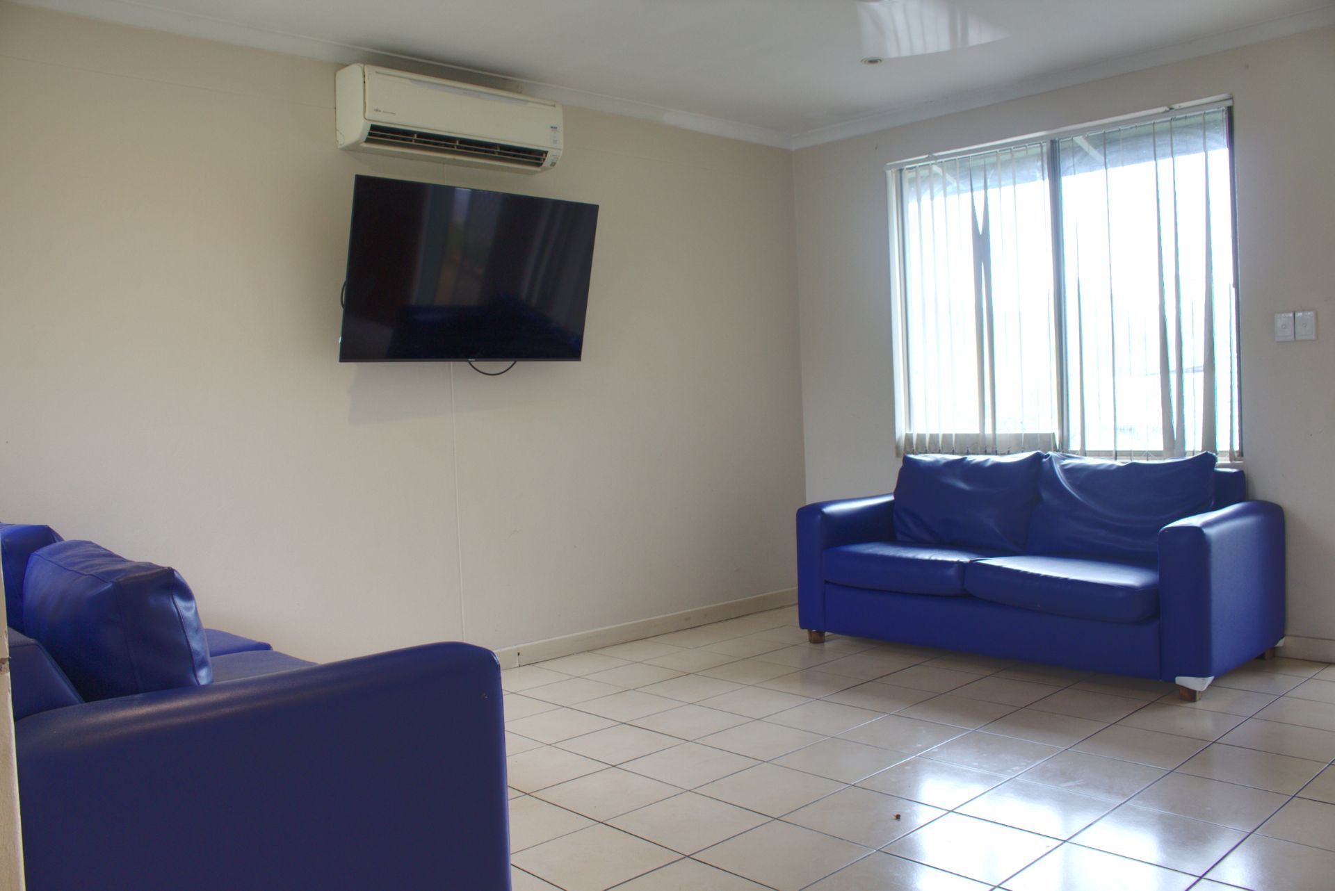 A single-story tan house with a green roof, a covered carport, and a small front garden with bushes on a paved driveway.