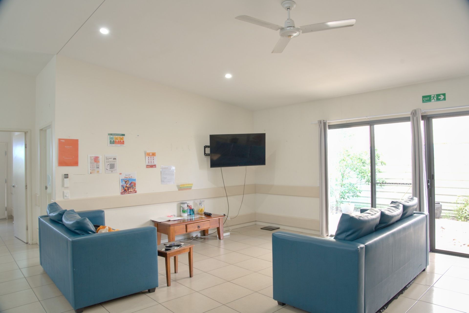 A modern lounge with two blue sofas, a wall-mounted television, a wooden coffee table, and large glass doors.