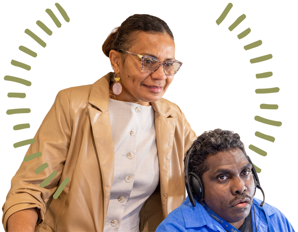 A person in a tan blazer stands behind a person wearing headphones and a blue shirt, both looking toward a screen.