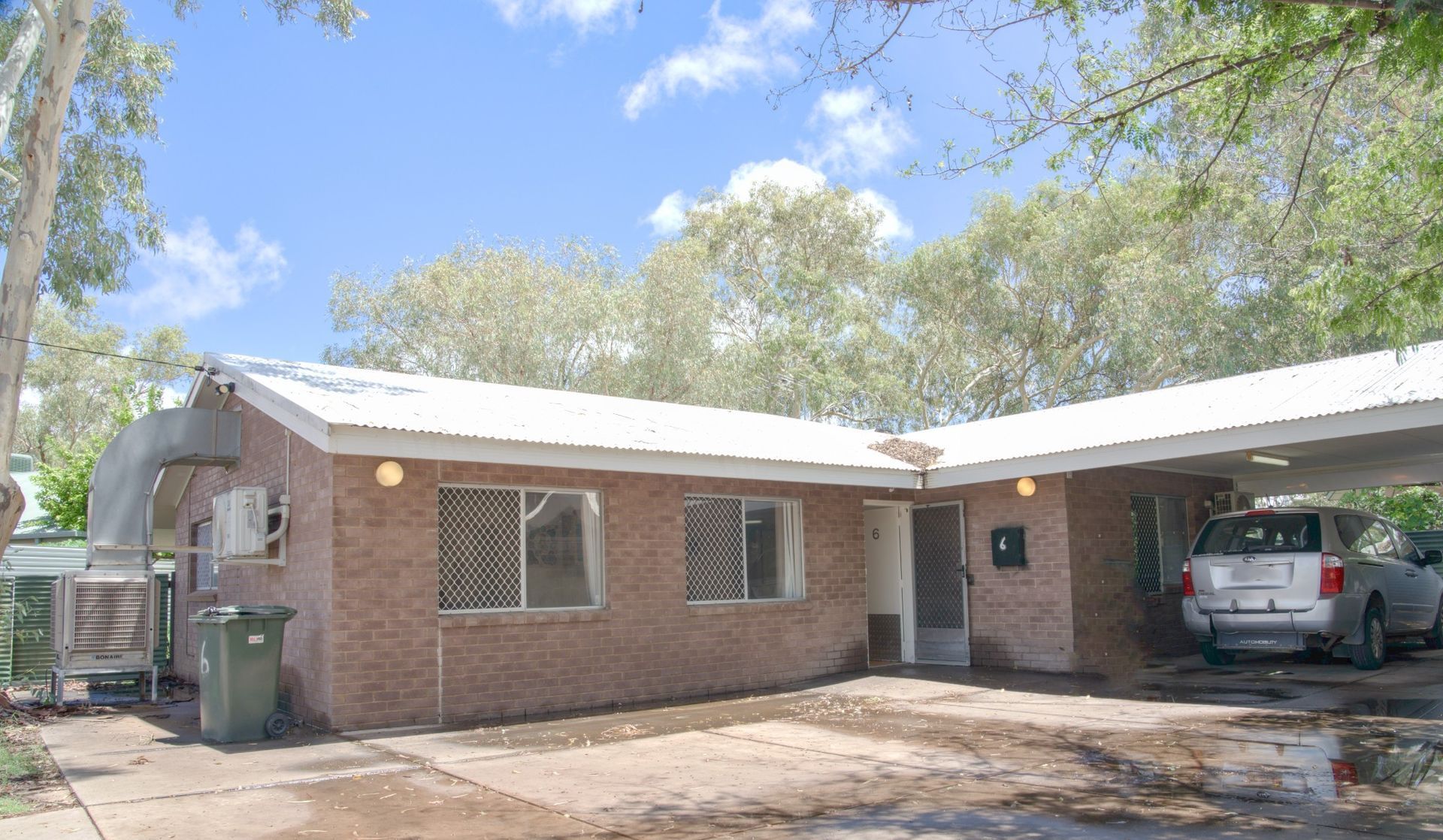 A single-story brick house with a white roof, a side carport with a silver SUV, and an outdoor cooling unit near the front.