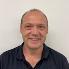 A person with short hair, wearing a dark-colored collared shirt, smiles warmly against a plain white background.