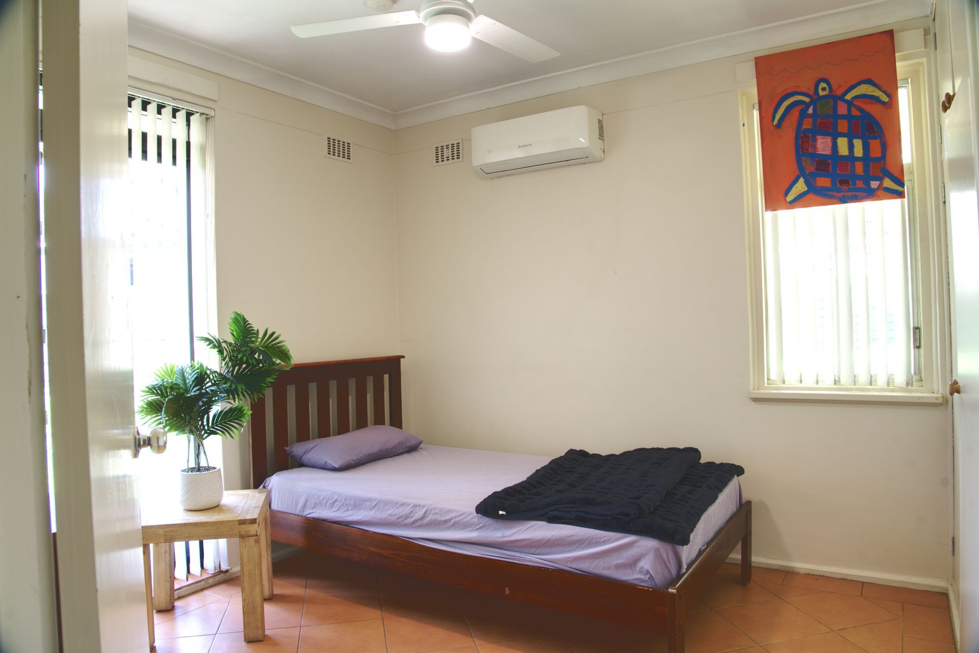 A bedroom with a wooden single bed, a small plant on a side table, and a turtle-patterned orange curtain on the window.