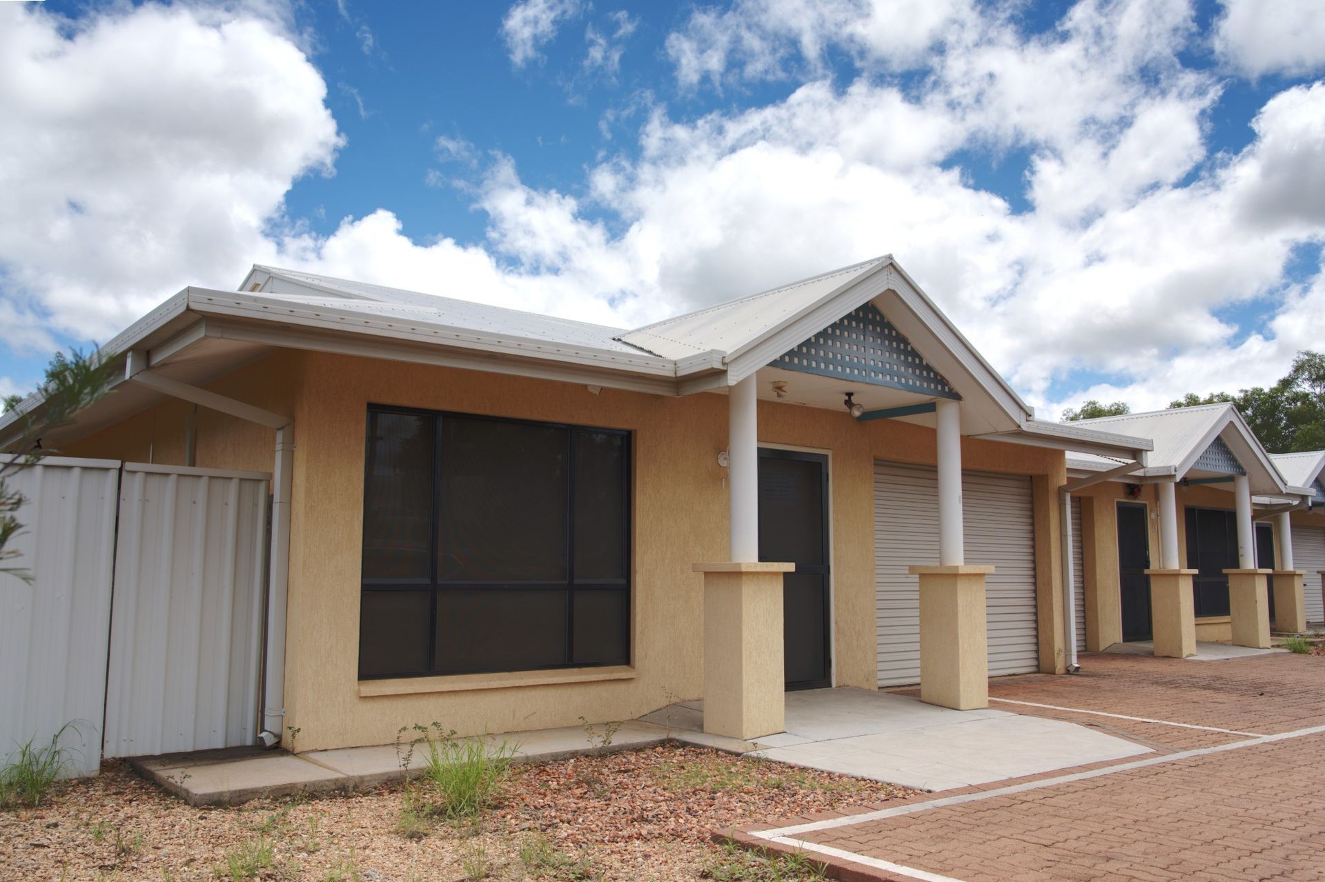 Single-story beige row houses with grey gabled roofs and pillars under a blue sky with white clouds.