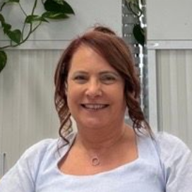A smiling person with auburn hair and a light blue top, posing in front of a plant and a wall partition.
