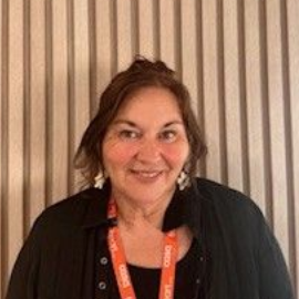 A person smiling in front of a vertical wood-slat wall, wearing a black shirt and an orange branded lanyard.