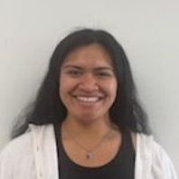A smiling person with long dark hair, wearing a white zip-up jacket over a dark top and a necklace against a white wall.