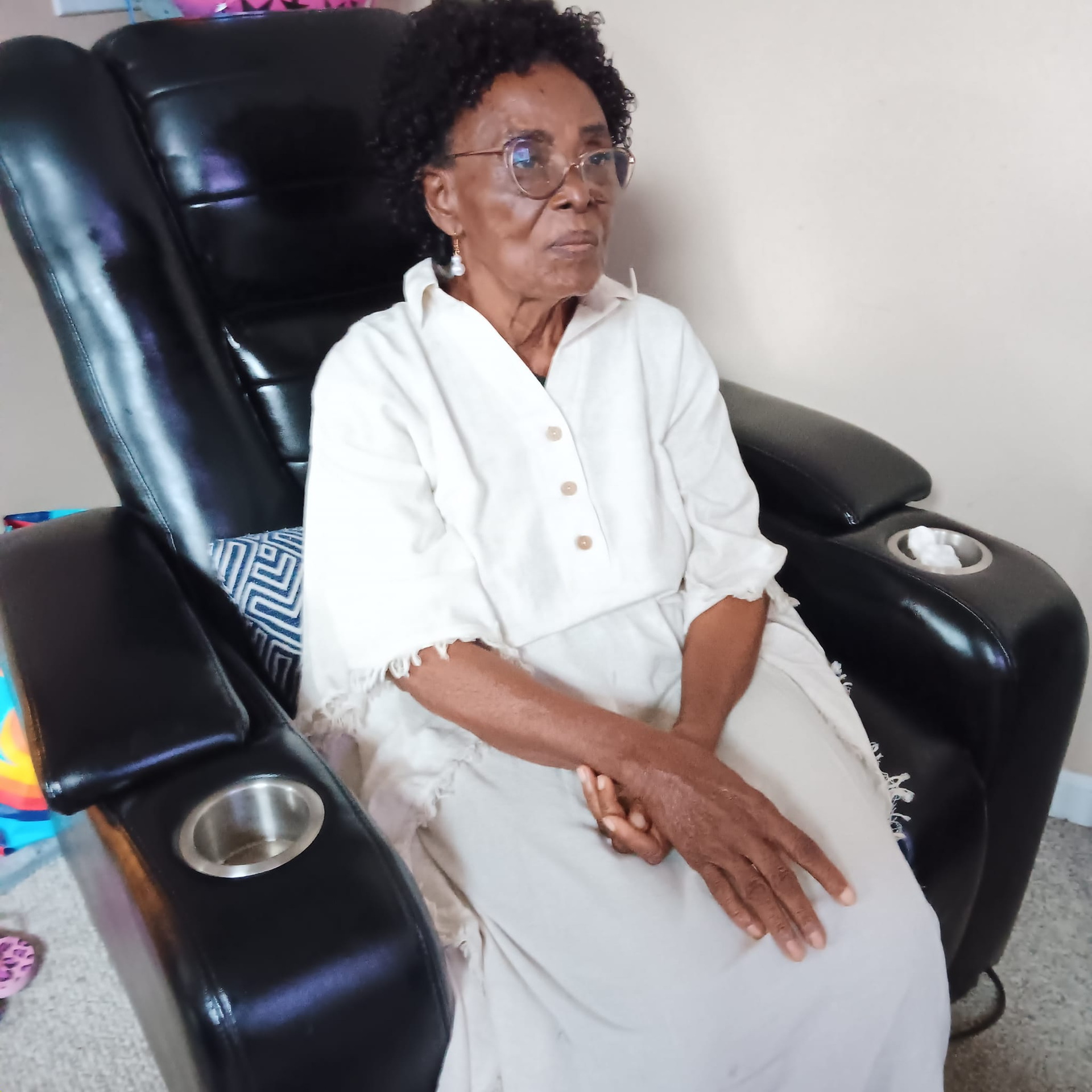 Woman sitting in a black massage chair, wearing glasses and a light-colored dress.