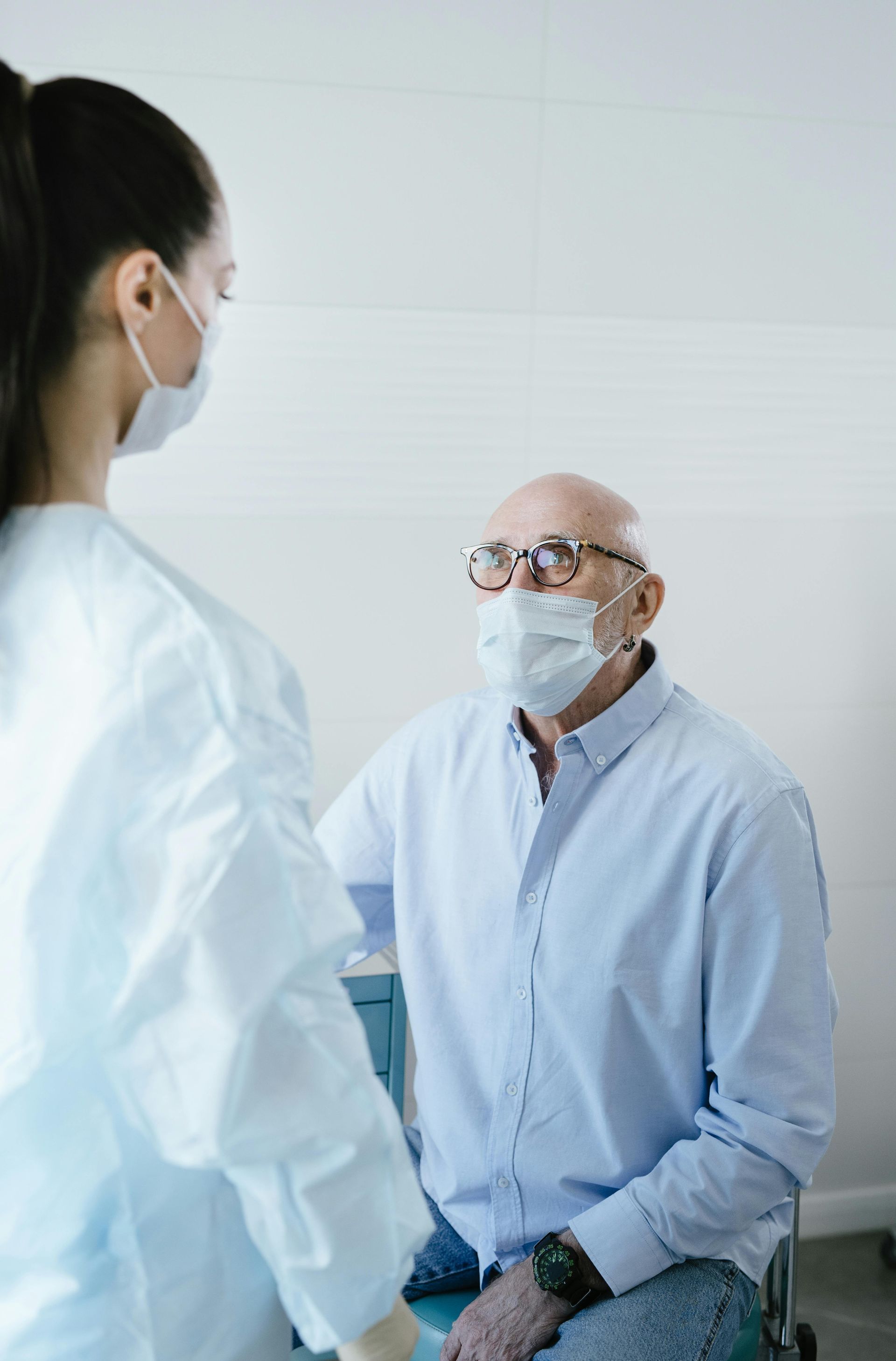 A medical professional in scrubs stands before a seated patient, both wearing face masks in a medical setting.