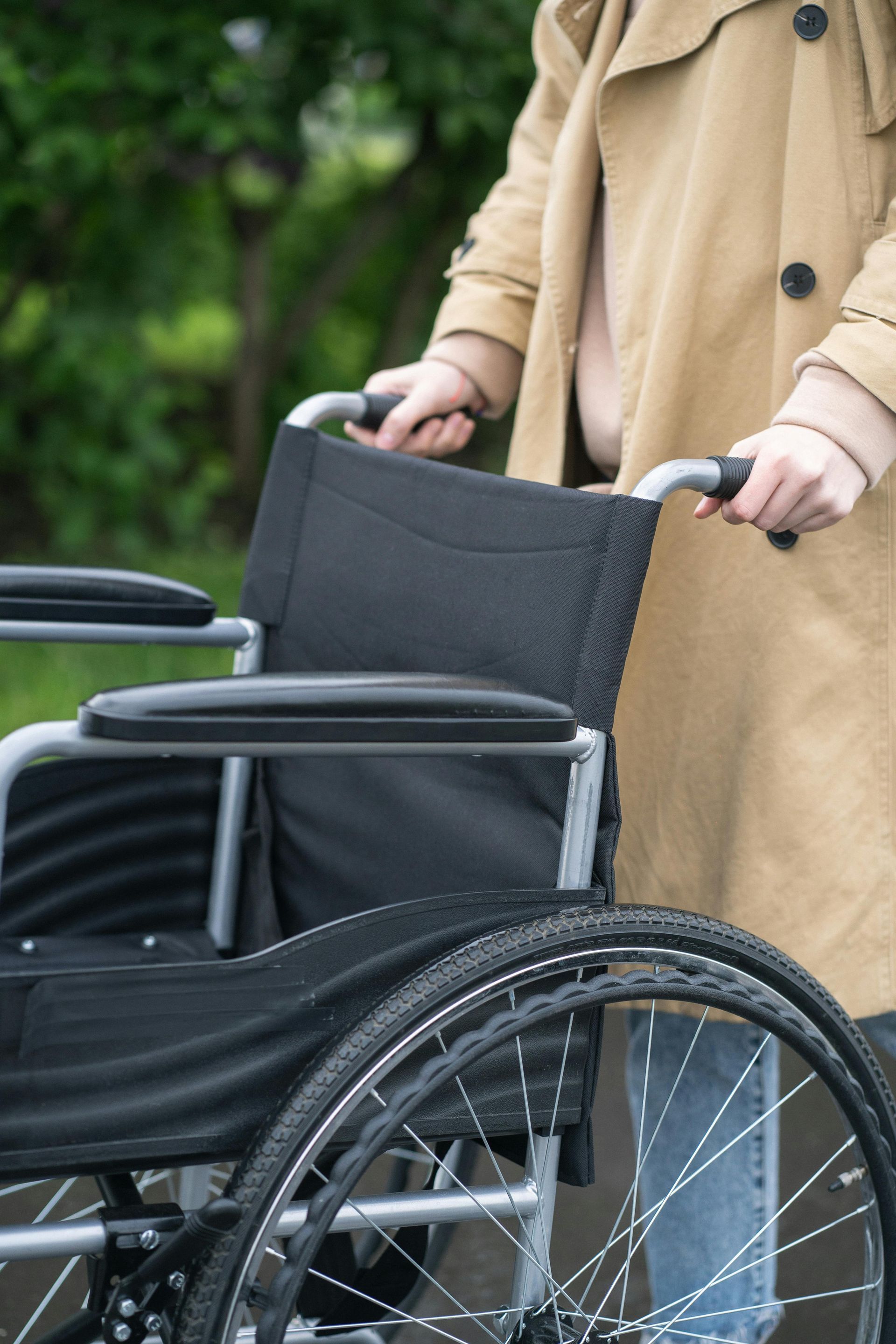 Person pushing a wheelchair outdoors. Beige coat, blue jeans. Black wheelchair.