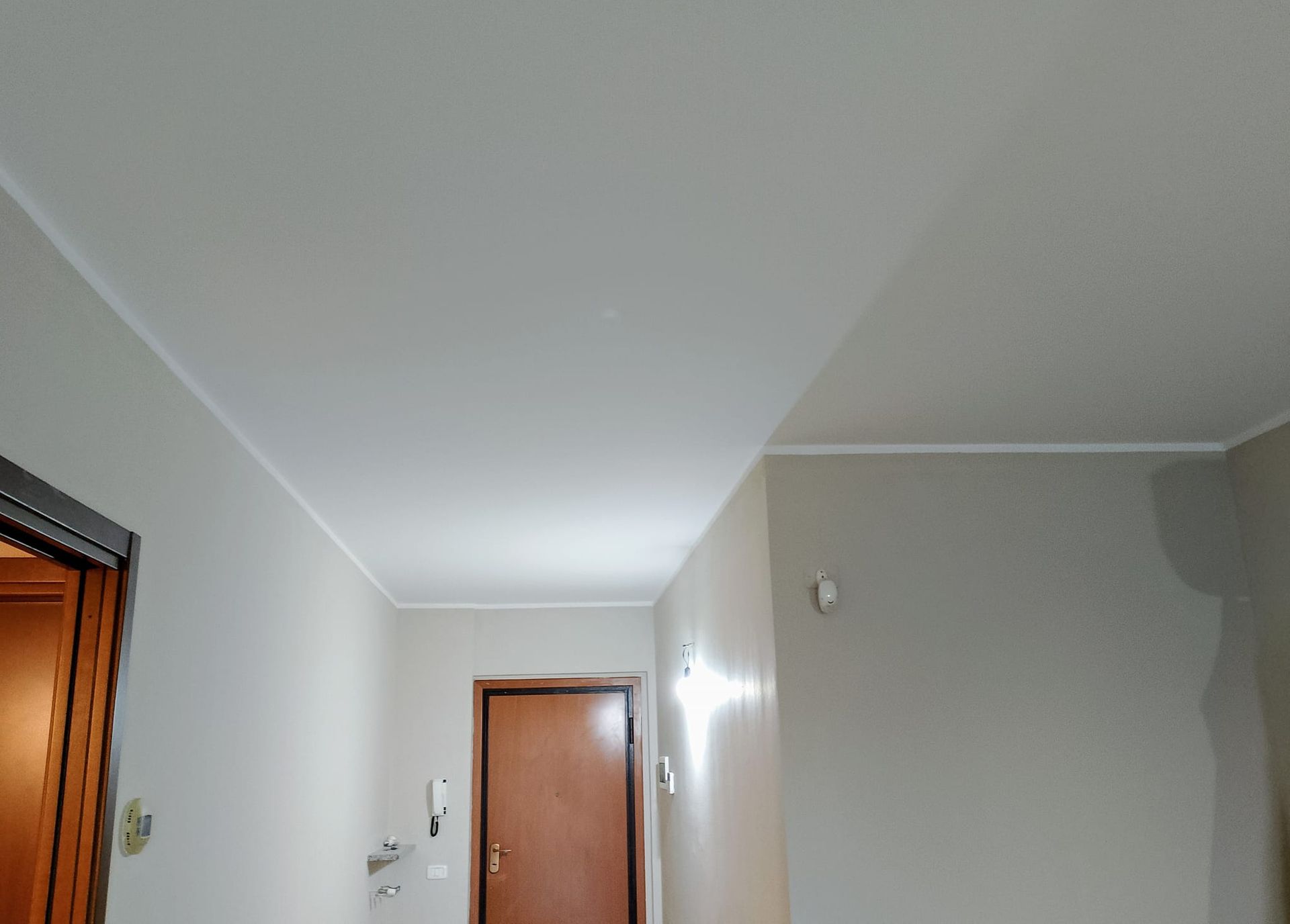 Un semplice corridoio interno con pareti e soffitto bianchi, una porta di legno marrone in fondo e un telaio di porta parziale sulla sinistra.