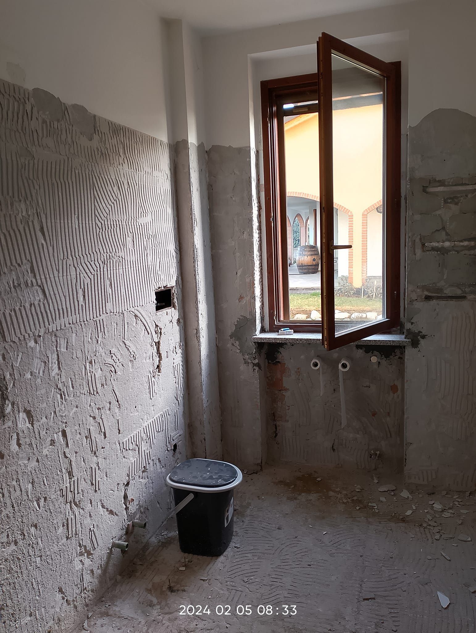 Un cantiere edile mostra una parete con adesivo steso a spatola, un secchio scuro sul pavimento e una finestra di legno aperta.