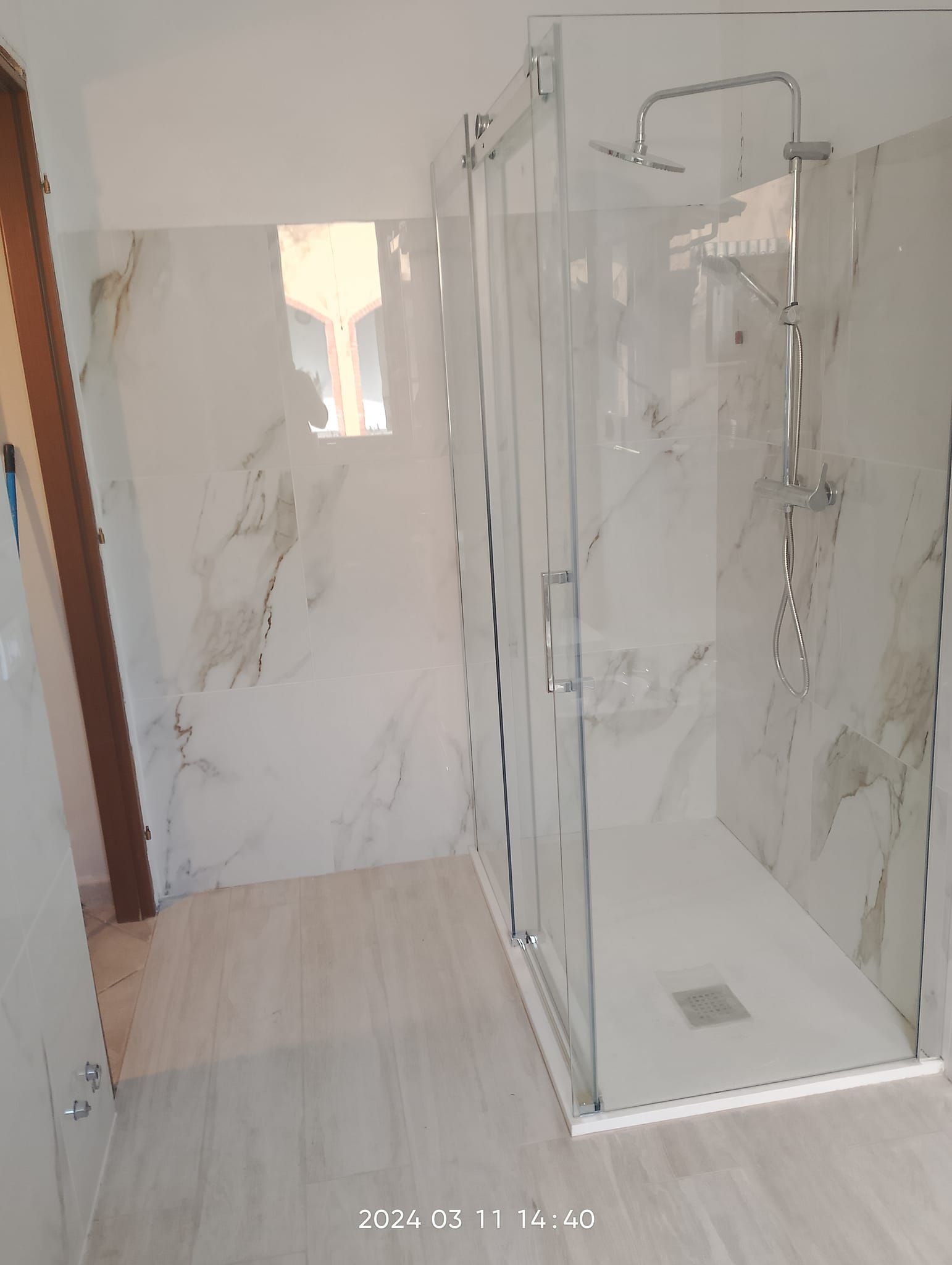 Un bagno moderno con doccia a box in vetro, pareti rivestite con piastrelle effetto marmo bianche e grigie e pavimento effetto legno chiaro.
