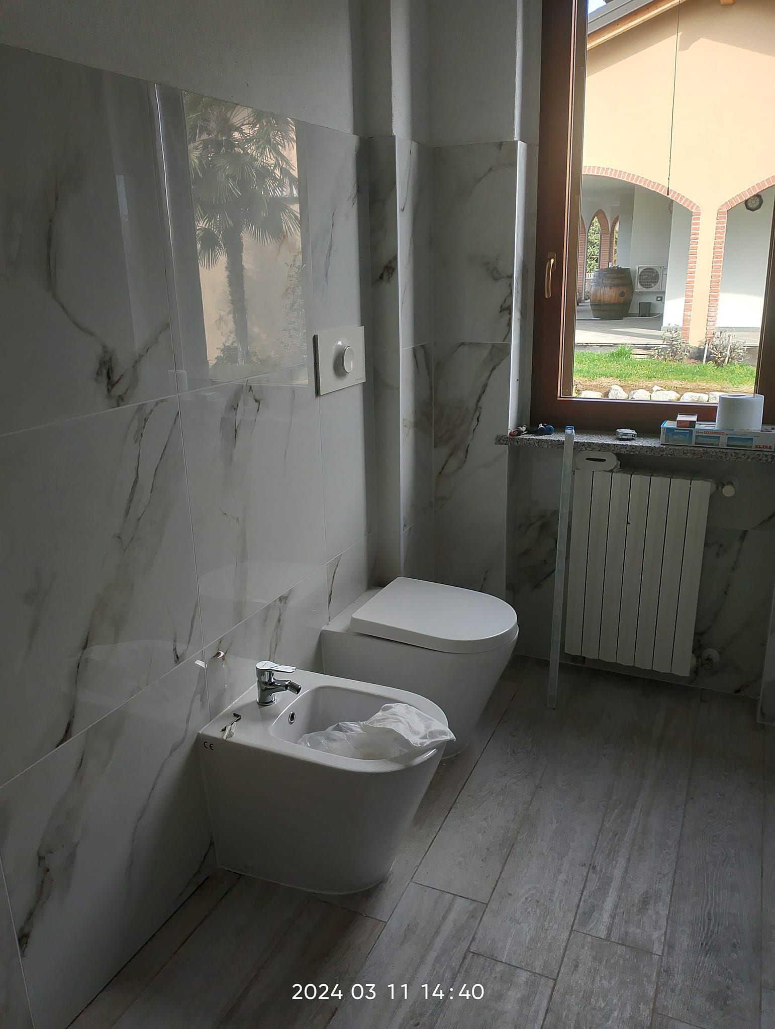 Un bagno moderno con bidet e WC bianchi addossati a una parete rivestita di piastrelle di marmo, vicino a una finestra con vista sull'esterno.