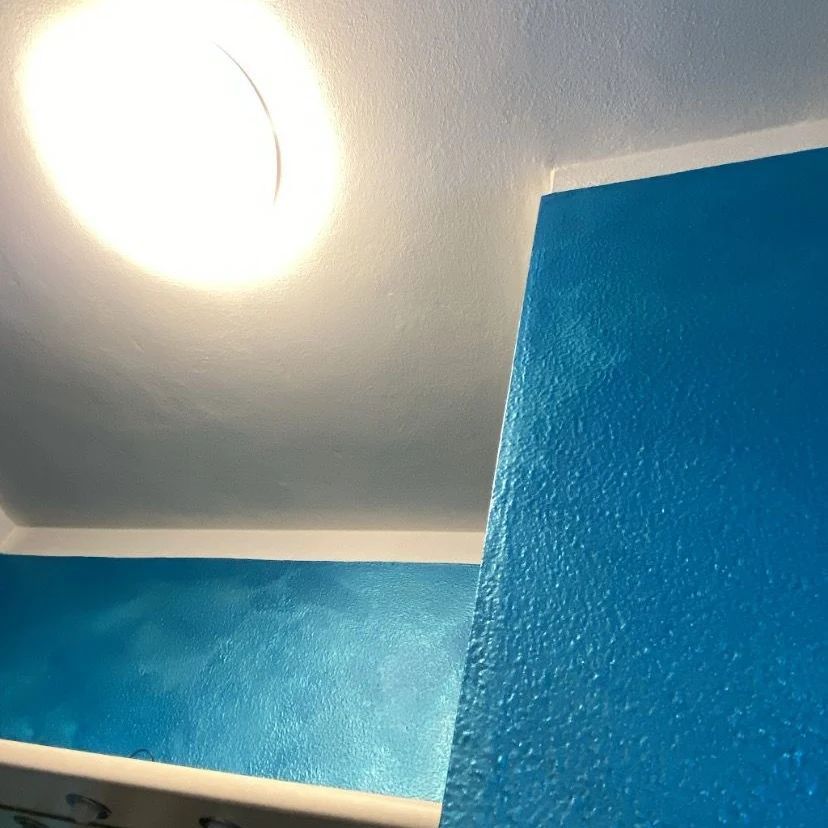 Una luminosa plafoniera circolare illumina un angolo dove una parete blu brillante e texturizzata incontra una parete e un soffitto bianchi.