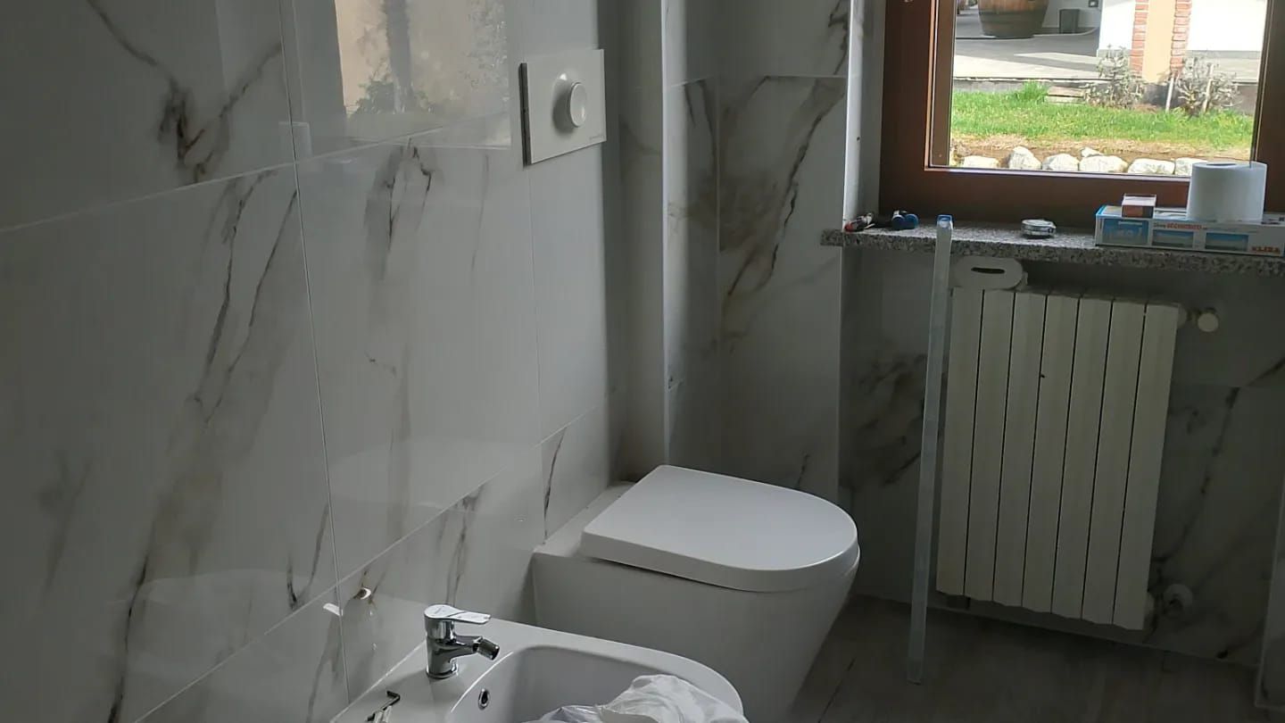 Un bagno moderno con bidet e WC bianchi, pareti rivestite con piastrelle effetto marmo chiaro e una finestra nelle vicinanze.
