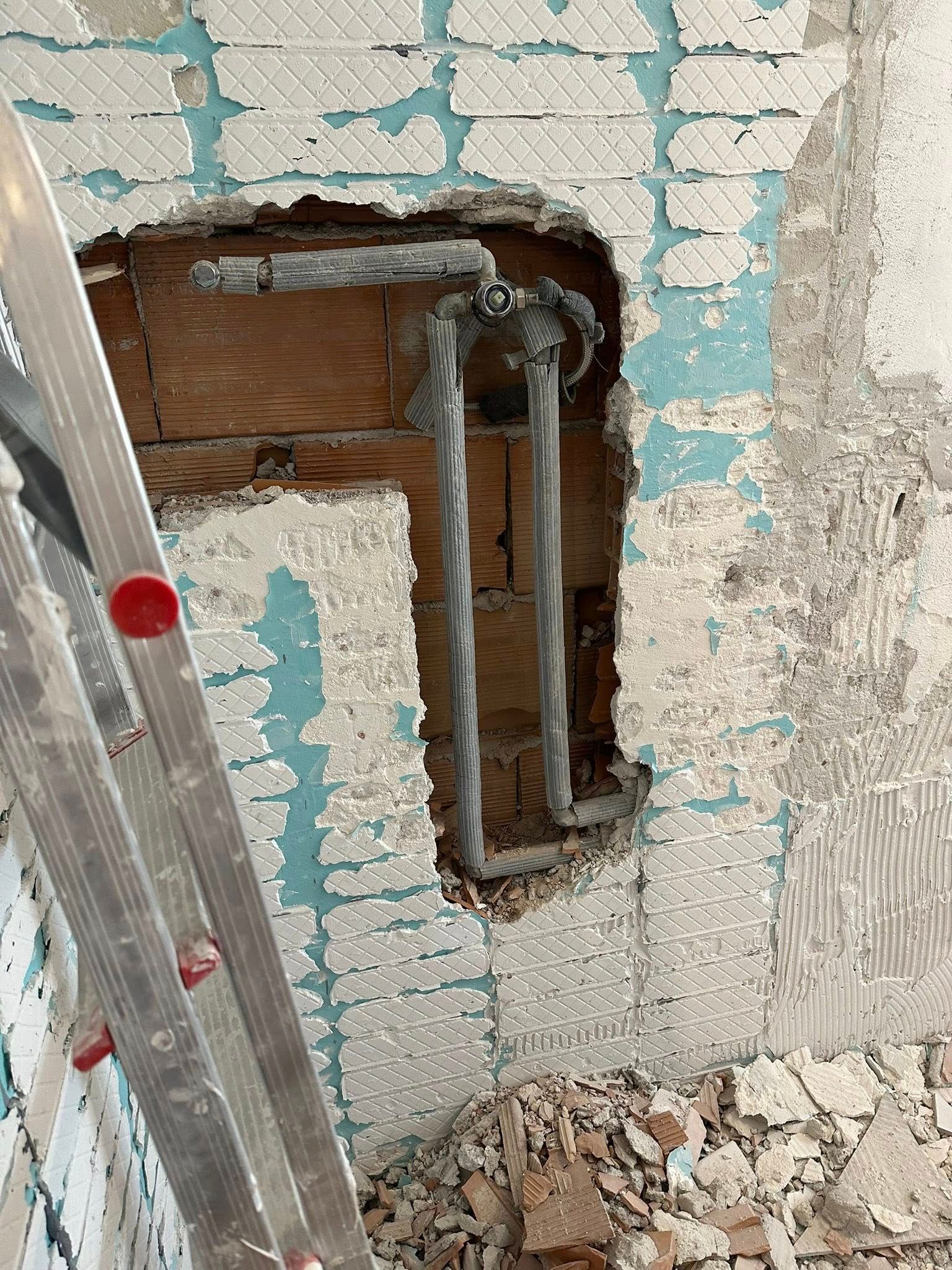 Una parete con una porzione di intonaco rimossa, che lascia intravedere tubi metallici grigi installati su blocchi di muratura di colore bruno-rossastro.