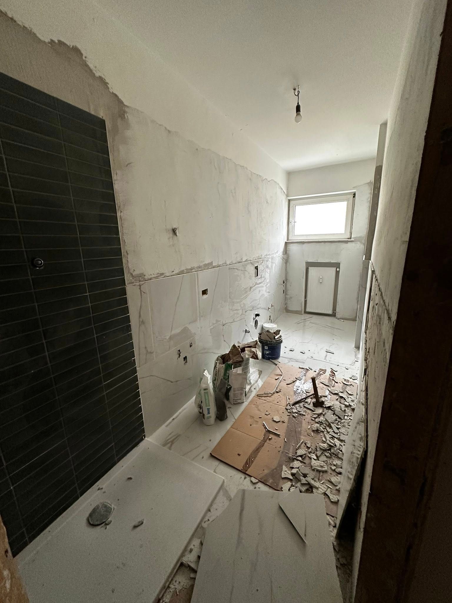 Una stanza in fase di ristrutturazione con una parete della doccia rivestita di piastrelle nere, pareti bianche non rifinite, detriti di costruzione e una finestra.