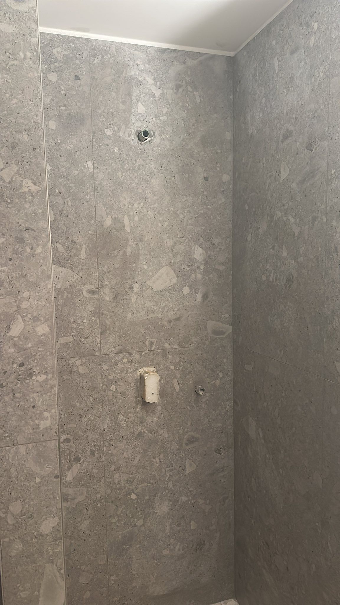 Una parete della doccia rivestita con piastrelle grigie effetto terrazzo, con miscelatore a parete e manopola di controllo separata.