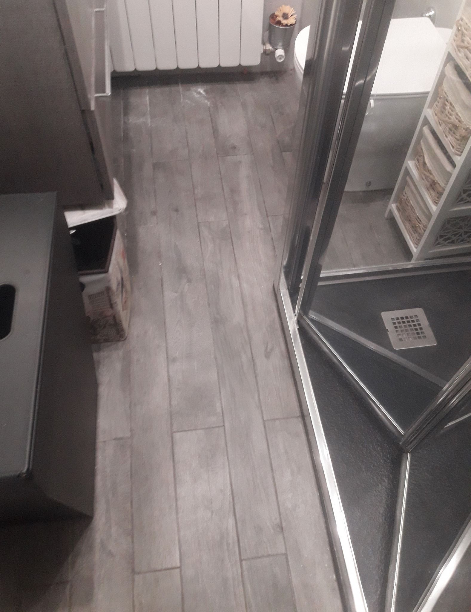 Un pavimento grigio effetto legno conduce alla zona bagno, dotata di termosifone bianco, WC bianco e scaffalatura in metallo argentato.