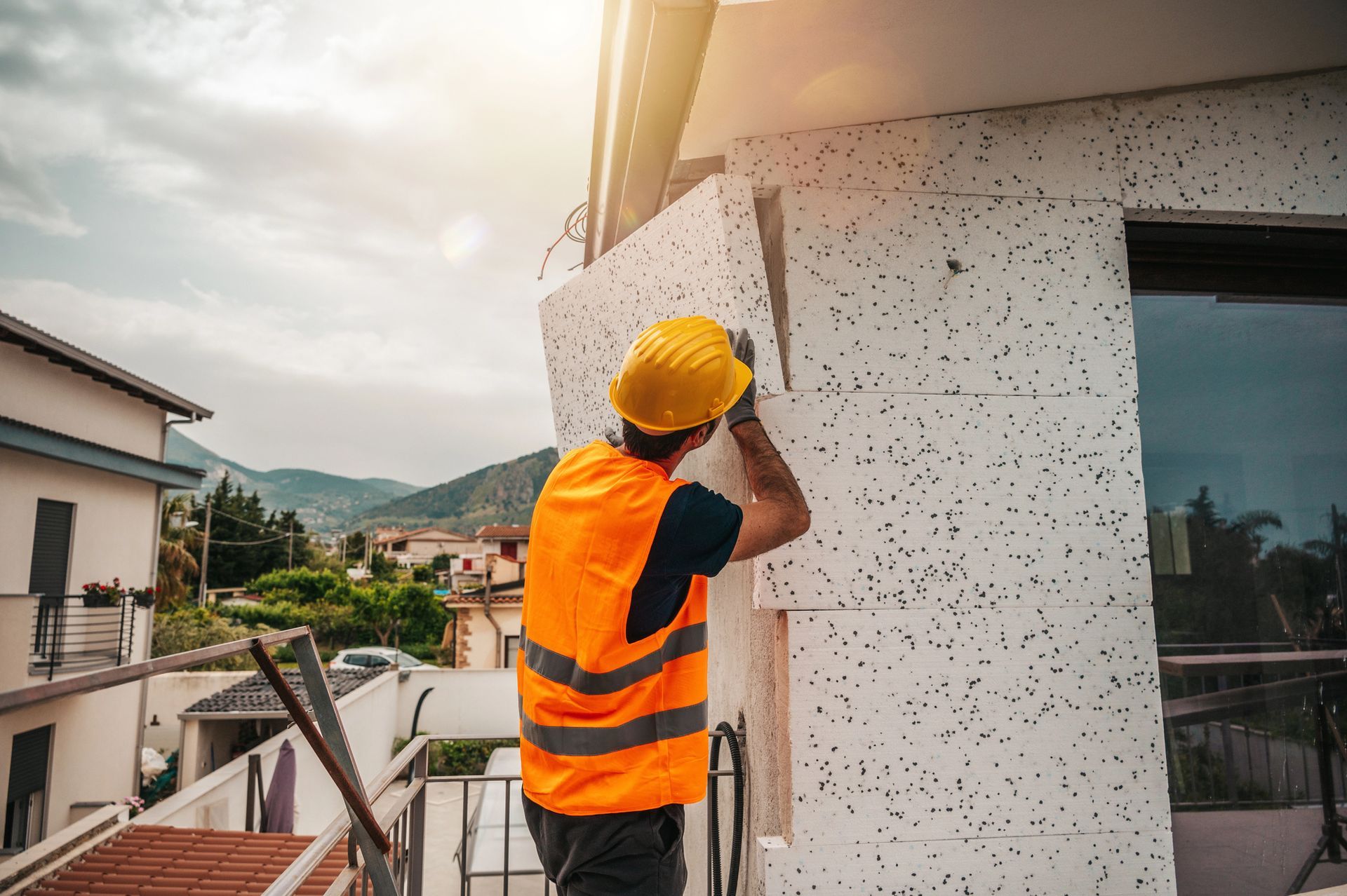 Un operaio edile con un elmetto giallo e un giubbotto di sicurezza arancione installa pannelli isolanti bianchi sulla facciata di un edificio.