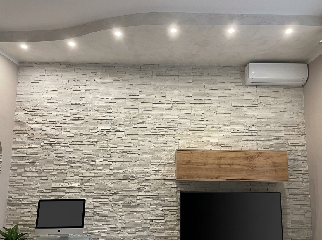 Una parete del soggiorno rivestita in pietra a vista, una mensola in legno sospesa sopra il televisore e faretti a incasso a soffitto.