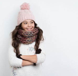 Niña con gorro, bufanda y suéter rosas, brazos cruzados, sonriendo.