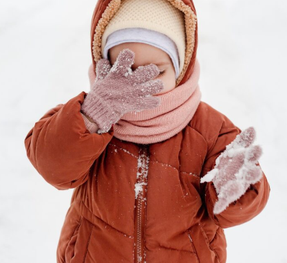 Niño con un abrigo naranja con capucha y bufanda y guantes rosas que le cubren la cara en la nieve.