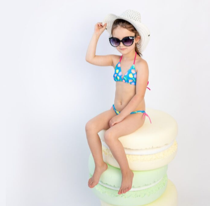 Una chica en bikini, gafas de sol y sombrero está sentada sobre una pila de macarons gigantes.