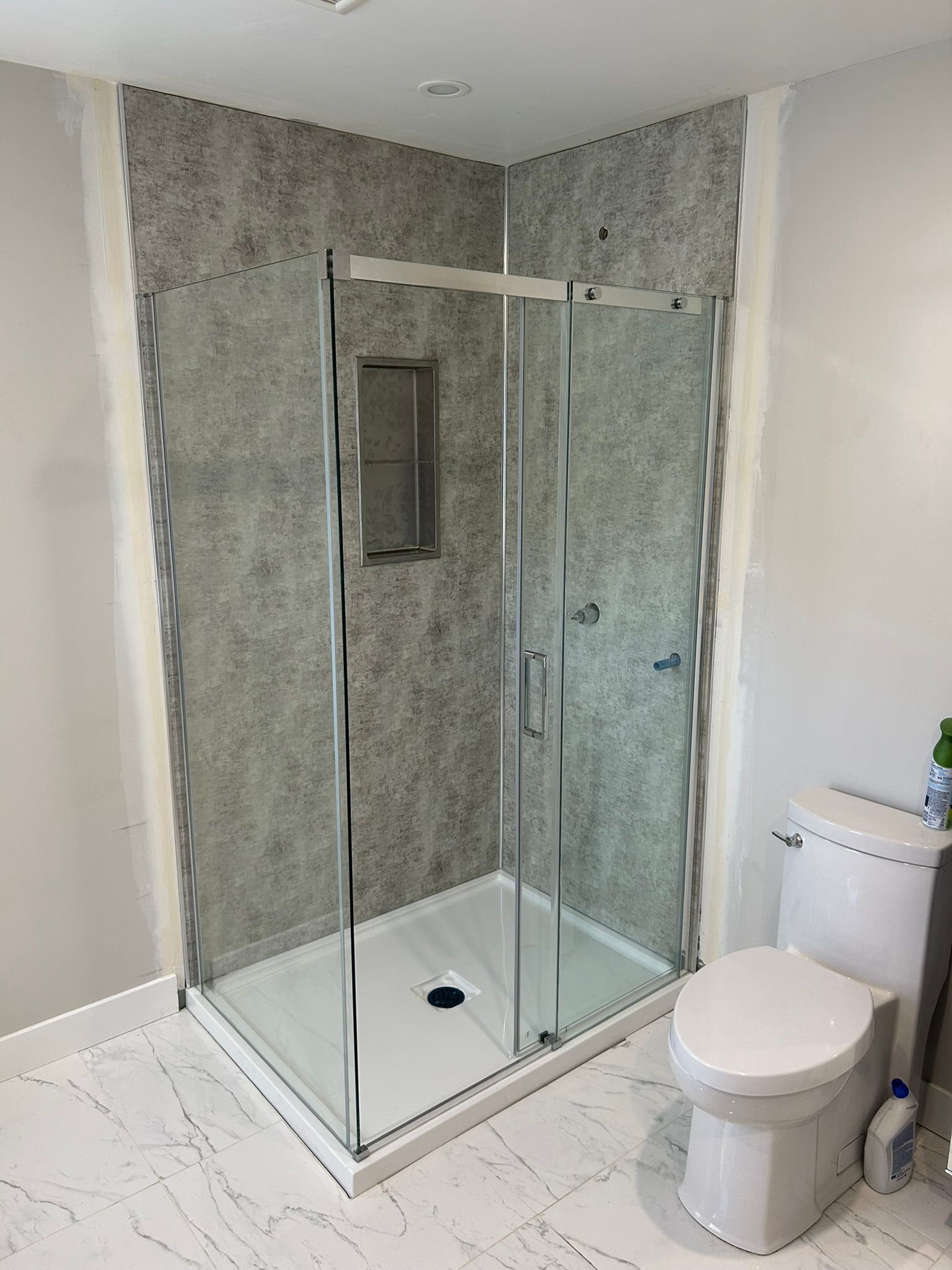 Une douche d'angle rénovée avec des murs gris et des portes en verre. Des toilettes blanches se trouvent à côté.