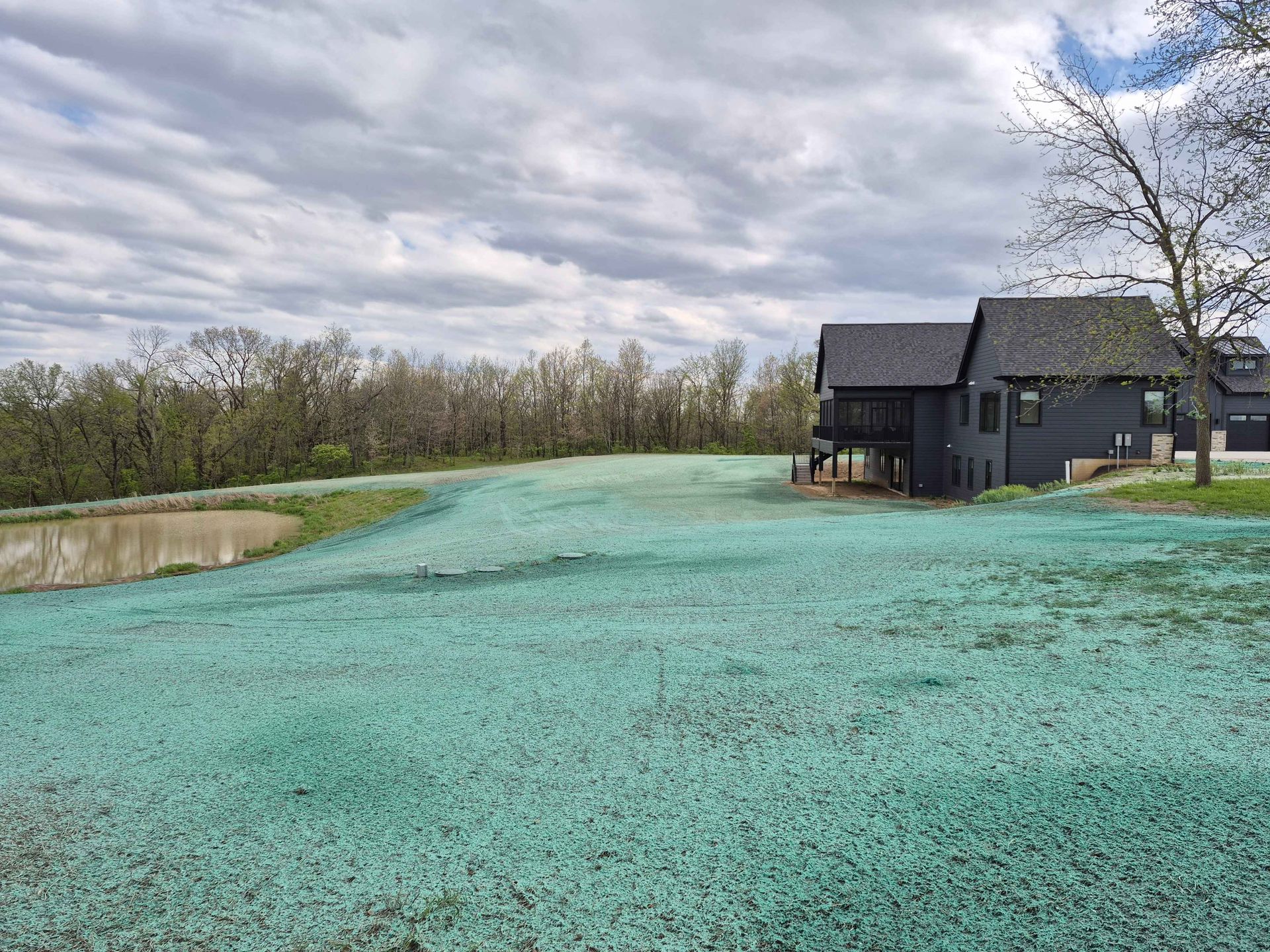 Dark house beside a turquoise pond and grassy field under a cloudy sky