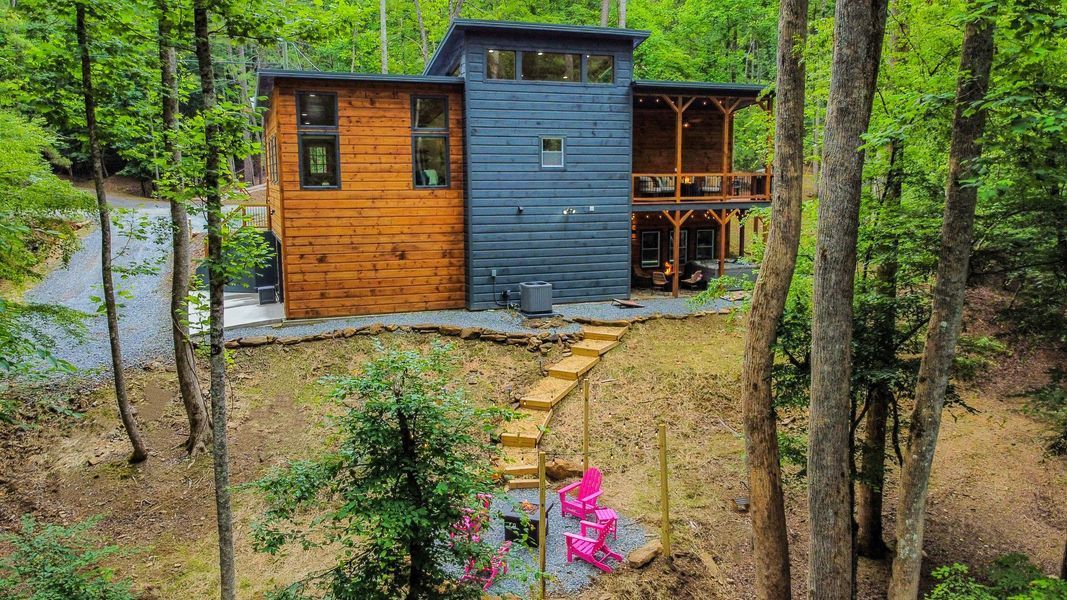 Modern house in a forest setting with wooden steps leading to a porch with pink chairs.