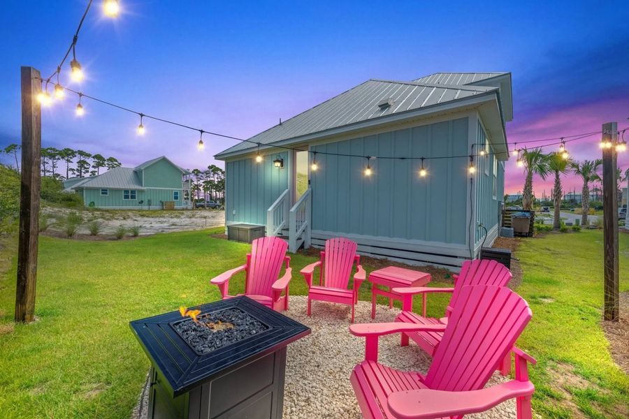 Backyard with pink chairs around a fire pit, lit by string lights. Blue house, green grass, dusk sky.