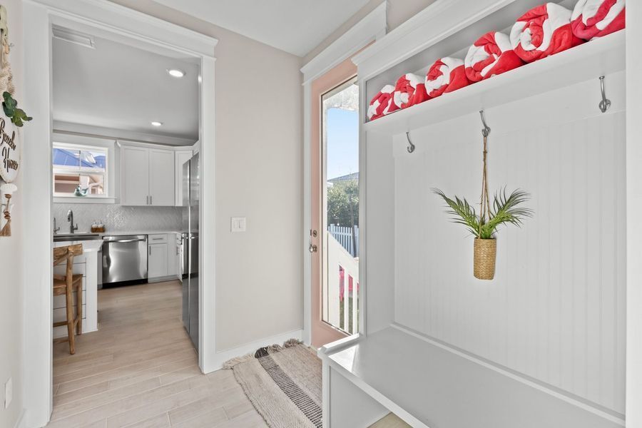 Entryway with white built-in bench, hooks, and shelves, pink door, leading to kitchen.