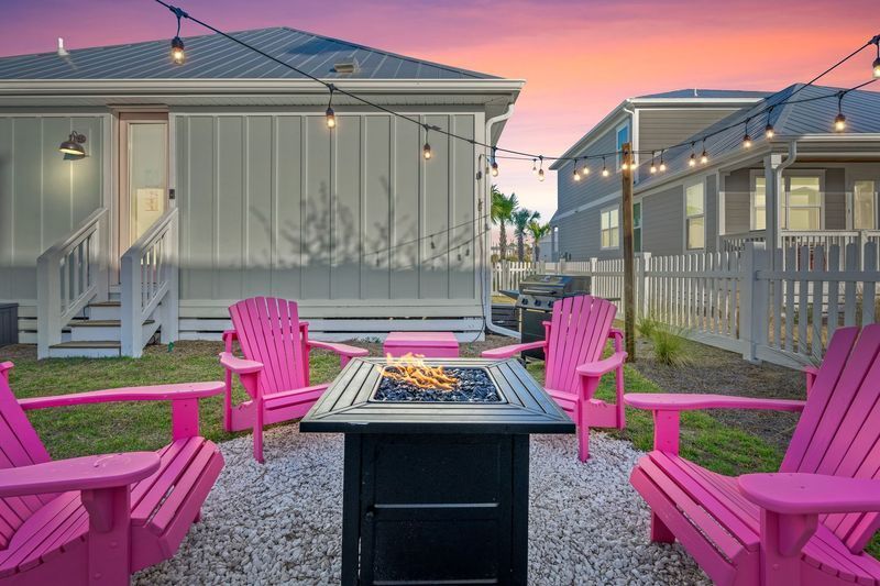 Backyard fire pit with pink chairs, surrounded by houses, lit by string lights against sunset.