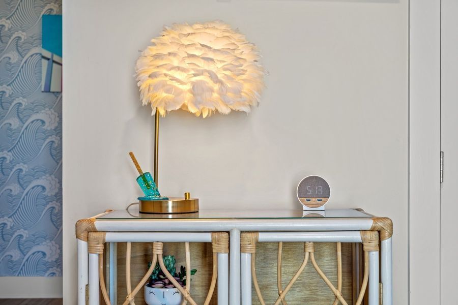 White feather lamp on a wicker table with decorative items against a white wall with blue wave wallpaper.