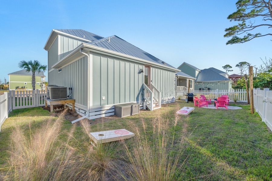 Backyard with a light blue house, white fence, and cornhole game set up. Pink chairs and grill are visible.