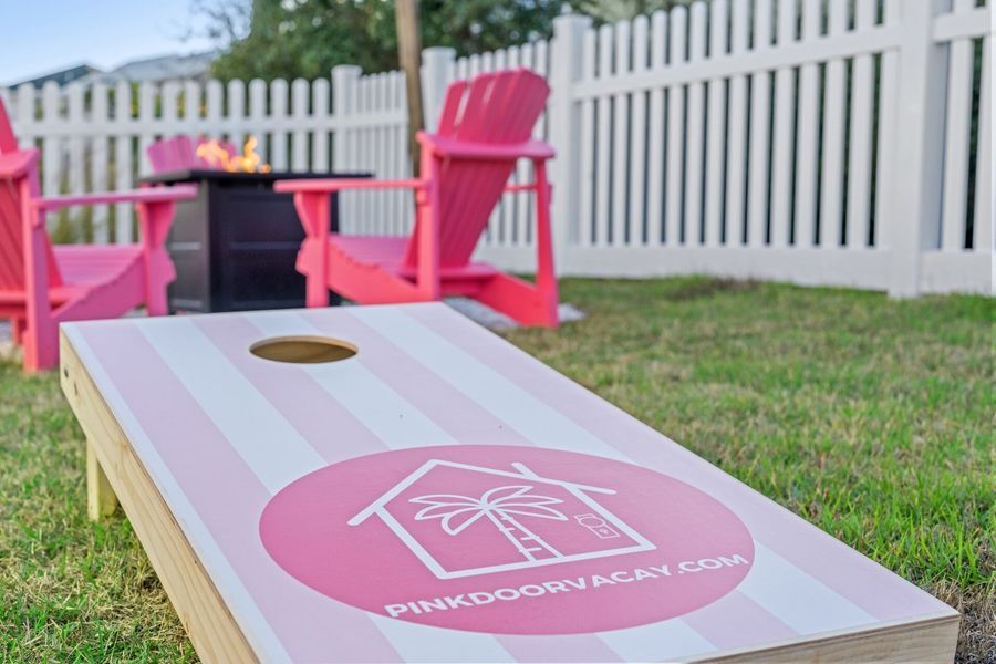 Cornhole board with pink logo, pink chairs, fire pit, and white picket fence in a grassy yard.