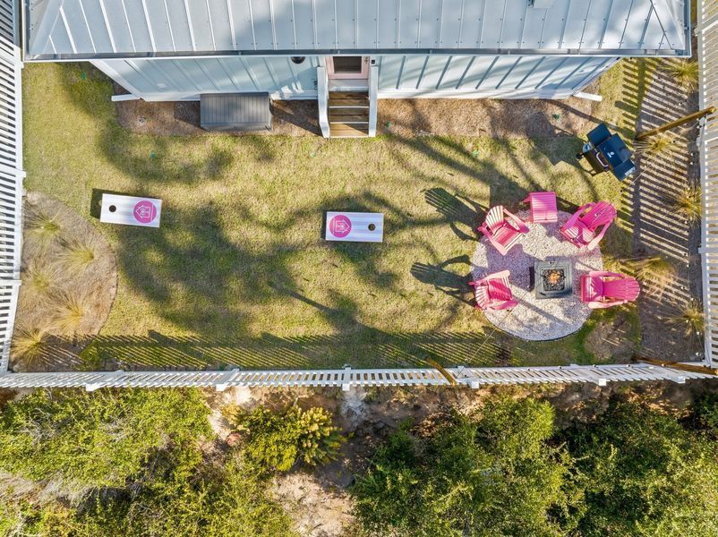 Overhead view of a backyard with a house, fire pit, cornhole, pink chairs, and white fence.