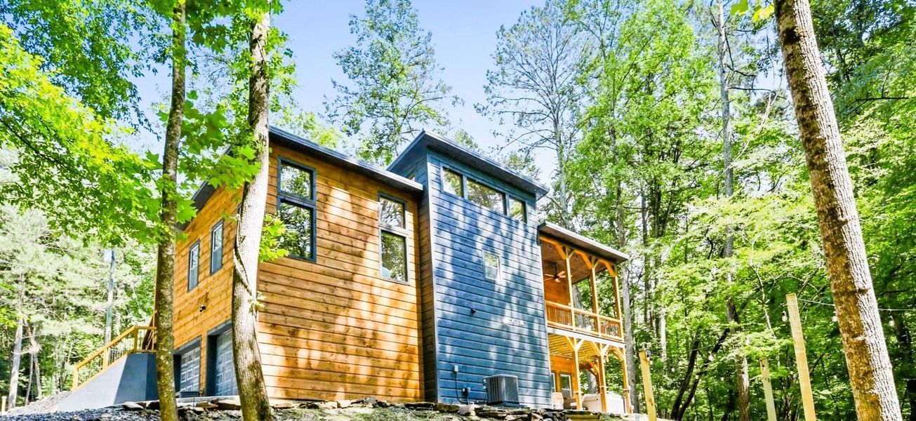 Cabin in the woods with wooden and blue exterior. Surrounded by lush green trees and blue sky.