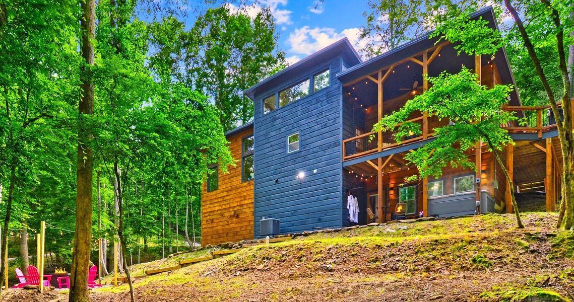 Modern two-story cabin with blue and wood siding, nestled in a green forest with a sunny sky.