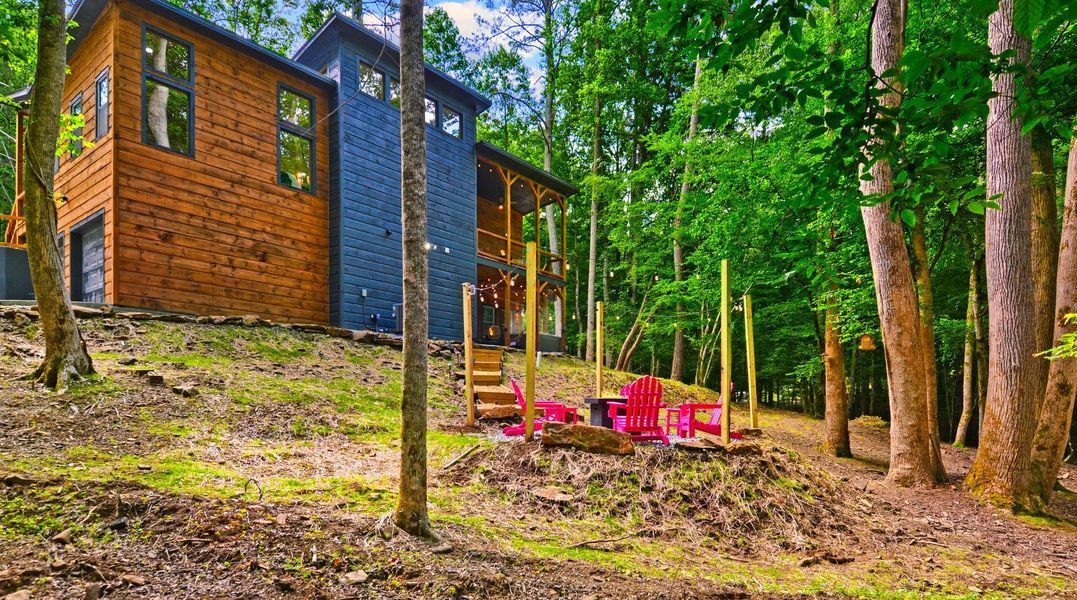 Cabin in a lush green forest; red Adirondack chairs near a small fire pit.