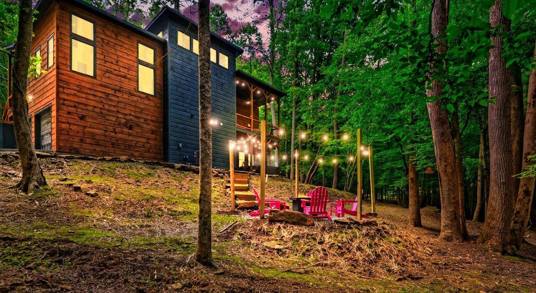 Cabin in the woods with string lights and red chairs in front of a fire pit.